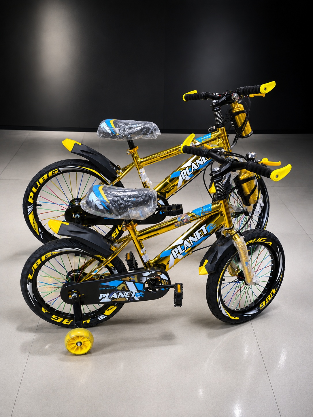 Two gold-and-black PLANET kids’ bicycles with training wheels, yellow accents, front suspension, and adjustable handlebars, displayed together on a light tiled floor against a dark
